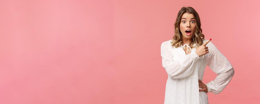 Surprised beautiful blond girl pointing upper right corner, gasping amused and staring at camera with fascinated expression, open mouth in awe to ask question about promo, pink background.