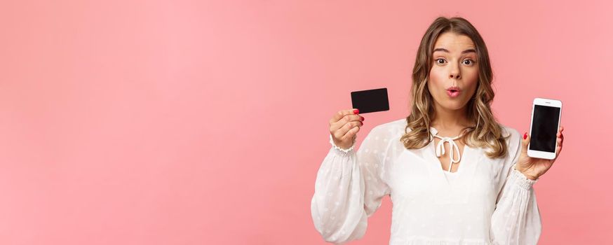 Finance, shopping and technology concept. Close-up portrait of excited blond cute girl in white dress, fold lips amused look camera as showing credit card and mobile phone, pink background.