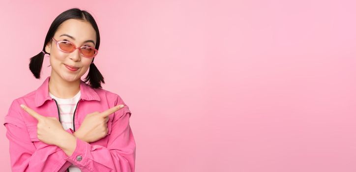 Choice. Stylish korean girl, asian female model points fingers sideways, shows two variants, product advertisement, demonstrating items, standing over pink background.