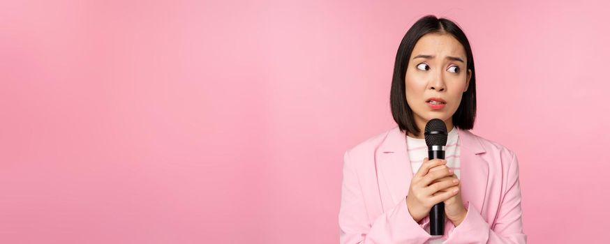 Insecure asian businesswoman giving speech, scared of talking in public, using microphone, standing in suit over pink background.