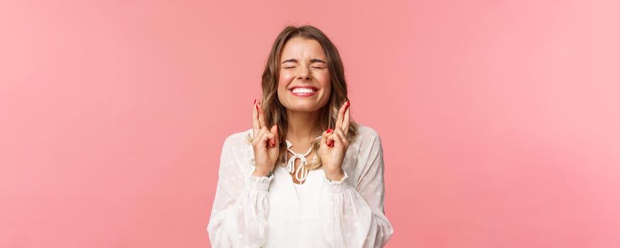 Portrait of excited hopeful blond girl making wish crossed fingers for good luck, close eyes and smiling putting all effort into pray, pleading for dream come true, anticipating over pink background.