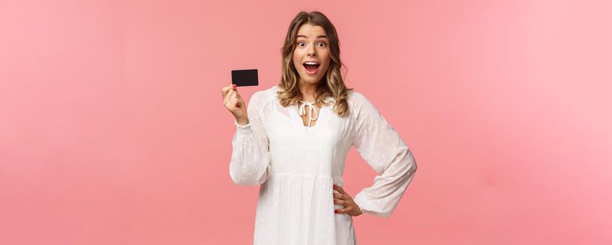 Portrait of happy excited good-looking blond girl in white dress, being impressed and pleased with cool banking service, showing credit card, smiling camera, standing pink background.