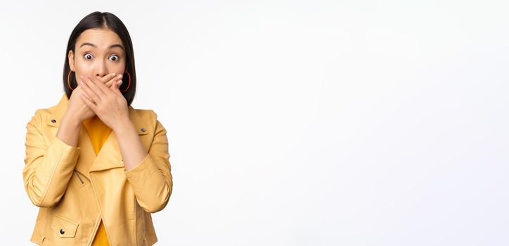 Image of shocked asian girl shuts mouth, close lips and looking speechless, startled face expression, standing over white background.