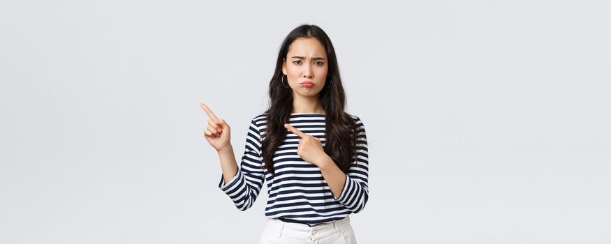 Lifestyle, beauty and fashion, people emotions concept. Disappointed sad asian girl complaining, feeling regret or jealousy as pouting uneasy and pointing fingers left at promo banner.
