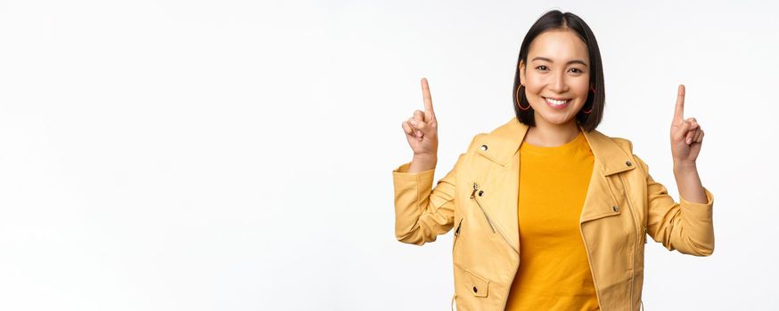 Enthusiastic asian girl pointing fingers up, showing advertisement on top, smiling happy, demonstrating promo offer or banner, standing over white background.