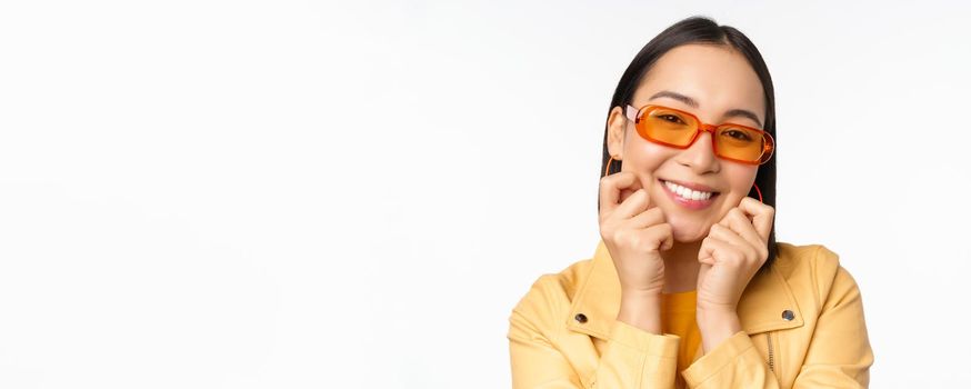 Close up portrait of trendy asian woman in sunglasses, touching her face, looking romantic, smiling at camera, standing over white background.