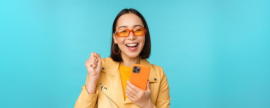 Happy stylish asian girl using smartphone and laughing, smiling at camera, standing over blue background. Copy space