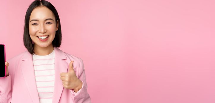 Satisfied smiling asian businesswoman recommending mobile phone app, website company on smartphone, showing screen and thumbs up, pink background.