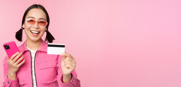 Modern beautiful asian girl, laughing and smiling with mobile phone, credit card, shopping online, paying with smartphone, standing over pink background.