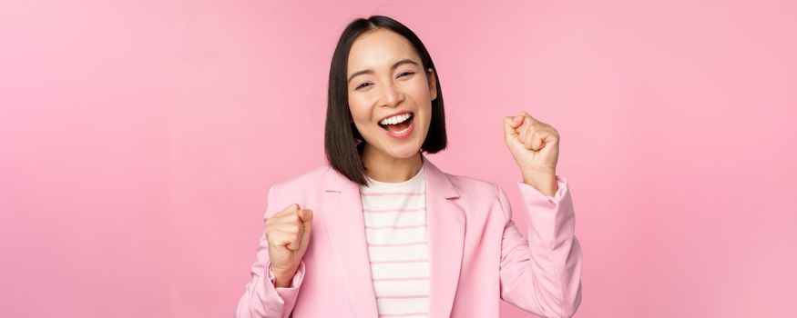 Enthusiastic saleswoman, asian corporate woman say yes, achieve goal and celebrating, triumphing, looking with rejoice and smiling, standing over pink background.