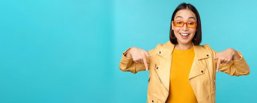 Portrait of stylish asian girl, wears sunglasses, smiles and points fingers down, shows advertisement, stands over blue background.