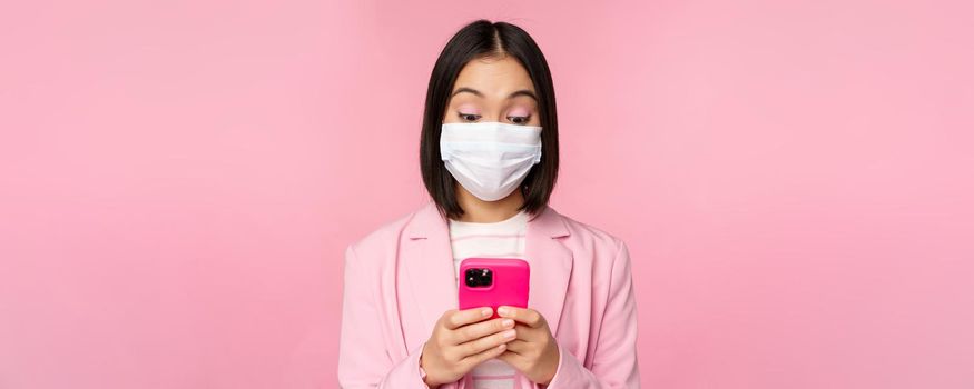 Business people and covid-19 concept. Young asian businesswoman in suit and medical face mask, using smartphone app, standing over pink background.