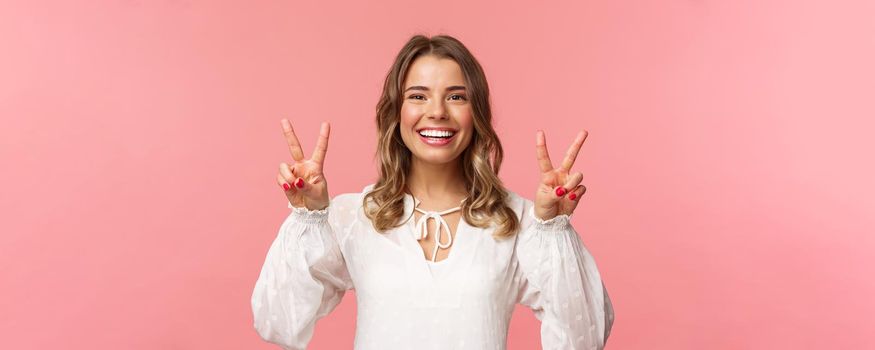 Close-up portrait of optimistic kawaii young blond girl with lovely beaming smile, showing peace signs and looking camera with positive attitude, enjoying spring, white dress, pink background.