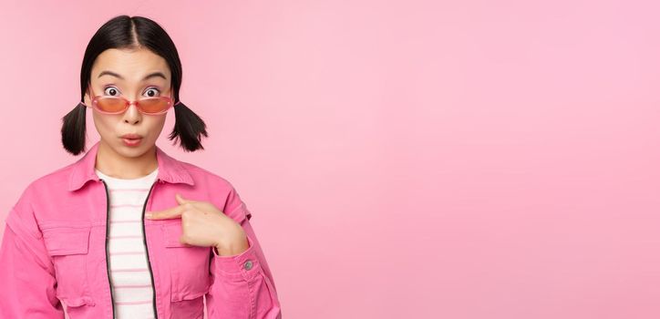 Close up portrait of asian girl looks surprised, points at herself with disbelief, being chosen, stands over pink background. Copy space