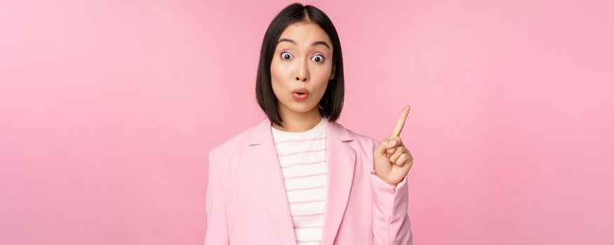 Got an idea. Young asian saleswoman, office manager raising finger, suggesting, wearing suit, posing against pink studio background.