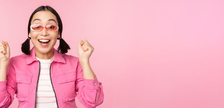 Portrait of excited japanese girl in sunglasses, celebrating, achieve goal, gasping amazed and smiling, standing over pink background.