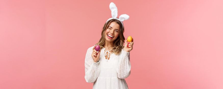 Holidays, spring and party concept. Cheerful good-looking blond woman celebrating Easter day in rabbit ears, holding two painted eggs and wink camera, smiling happily, pink background.