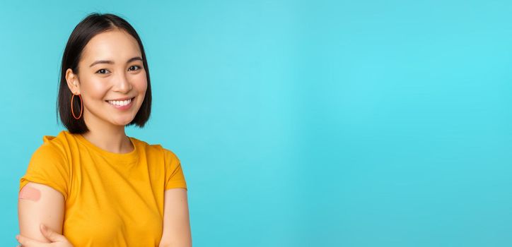 Vaccine campaign from covid-19. Young beautiful, healthy asian woman showing shoulder with bandaid, concept of vaccination, standing over blue background.