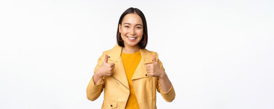 Cheerful asian woman smiling pleased, showing thumbs up in approval, standing in casual clothes over white background.