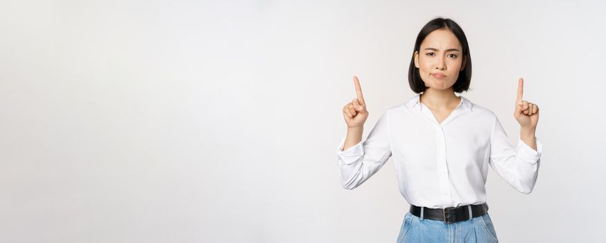 Image of skeptical asian lady grimacing, frowning upset, pointing fingers up, showing advertisement, standing over white background.
