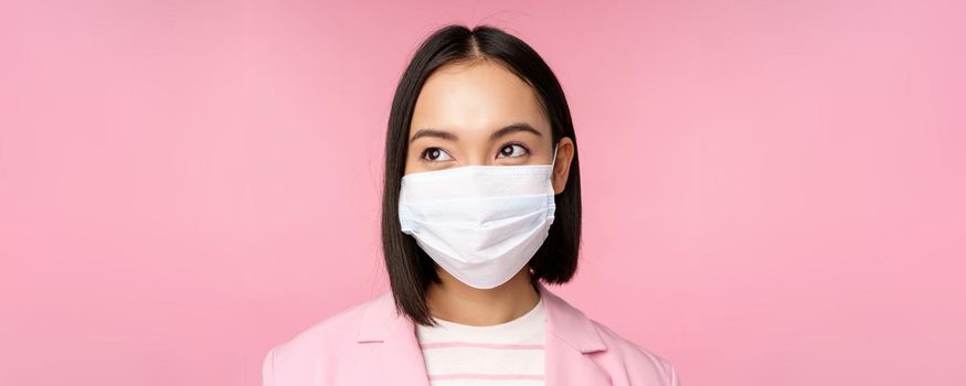 Close up portrait of japanese corporate woman in medical face mask from covid-19, looking left at logo, sale promo, standing over pink background.