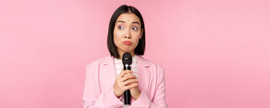 Nervous female entrepreneur giving speech with microphone, holding mic and looking aside anxiously, posing in suit over pink background.