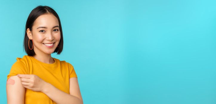 Vaccine campaign from covid-19. Young beautiful, healthy asian woman showing shoulder with bandaid, concept of vaccination, standing over blue background.