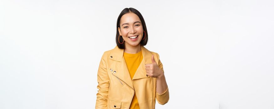 Beautiful korean girl smiling, showing thumbs up, like gesture, recommending store or company, standing satisfied against white background.