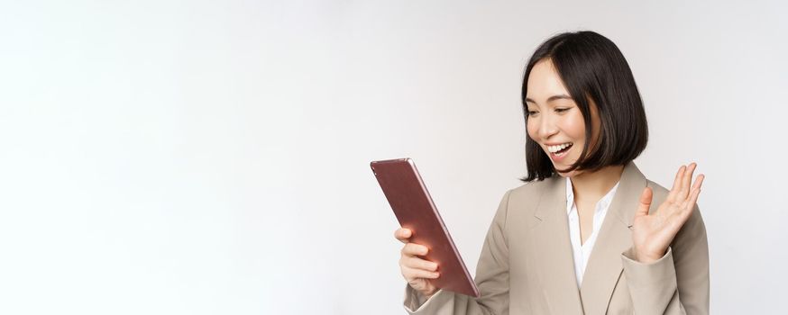 Portrait of smiling asian businesswoman video chat on digital tablet, waving hand at gadget screen, standing in suit over white background.