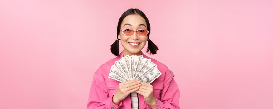Microcredit and fast loans concept. Excited stylish korean girl, showing money, cash dollars and looking happy, standing in sunglasses over pink background.