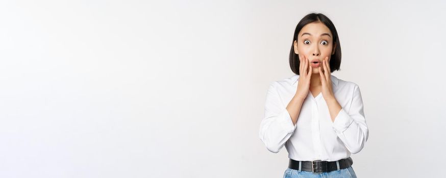 Image of asian female model looking surprised, staring amazed, reacting at surprise big news, standing over white background.