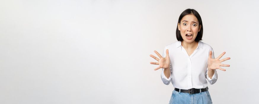 Asian woman looks at camera and screams in panic. Young korean girl looking anxious, panicking, shaking hands and shouting, standing over white background.