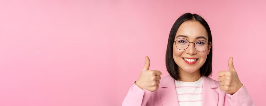Awesome, congrats. Face of excited asian businesswoman in glasses, smiling pleased, showing thumbs up in approval, standing over pink background.