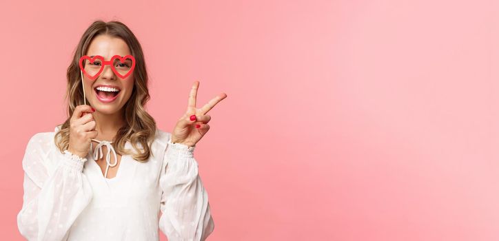 Spring, happiness and celebration concept. Close-up portrait of upbeat blond girl at party, wear white dress holding heart-shaped glasses mask over eyes and make peace sign cheerful.