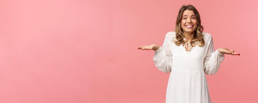 Beauty, fashion and women concept. Sorry dont know, cant say. Cheerful cute silly blond girl 20s, wear white dress, shrugging with hands sideways make awkward smile, apologizing, pink background.