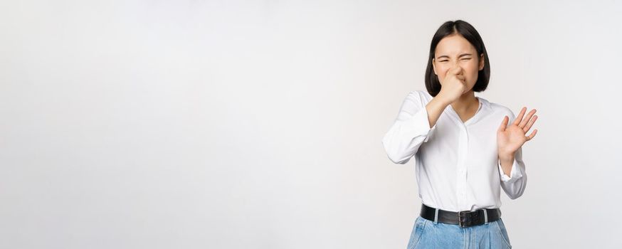 Disgusted asian girl close nose and showing rejection gesture, digusting bad smell, standing over white background.