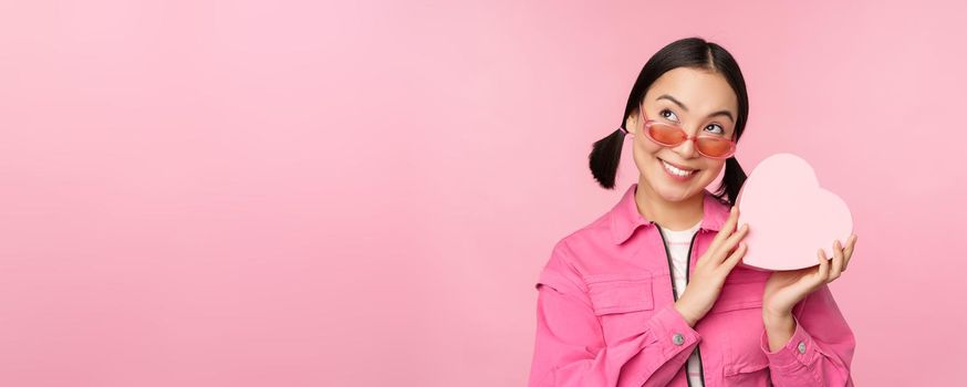 Image of stylish asian girlfriend in sunglasses, guessing whats inside gift box, heart-shaped present, standing over pink background.