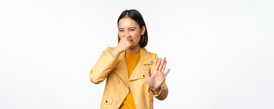 Image of asian girl shuts nose and reject smth with disgusting bad smell, shuts nose, standing over white background. Copy space