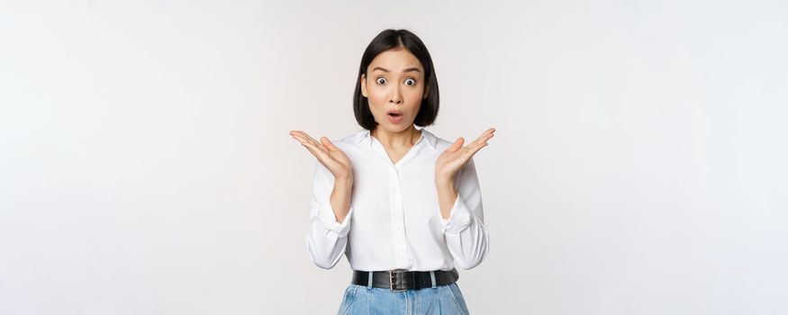 Image of asian female model looking surprised, staring amazed, reacting at surprise big news, standing over white background.