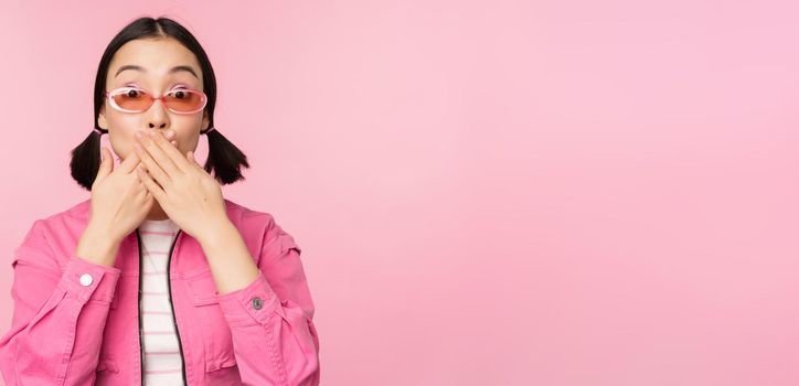 Portrait of shocked, stylish asian girl in sunglasses, closes mouth, looks with surprised face expression, stands over pink background. Copy space