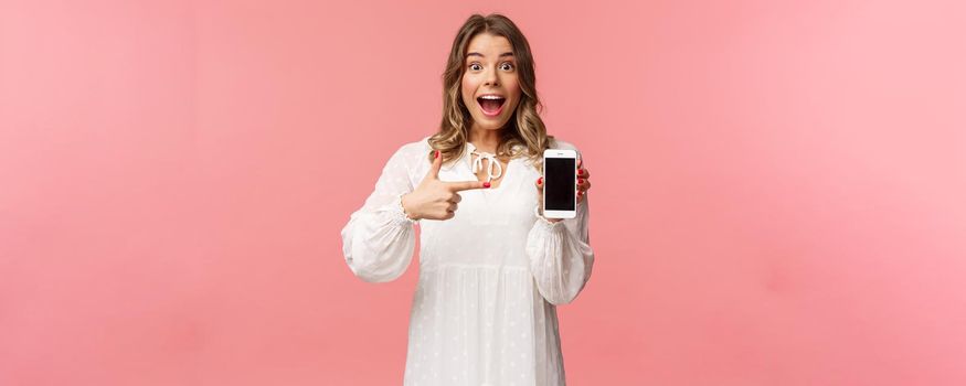 Portrait of impressed, excited young blond woman showing something awesome on display, pointing mobile phone screen and smiling astonished, brag with her recent match on dating app.