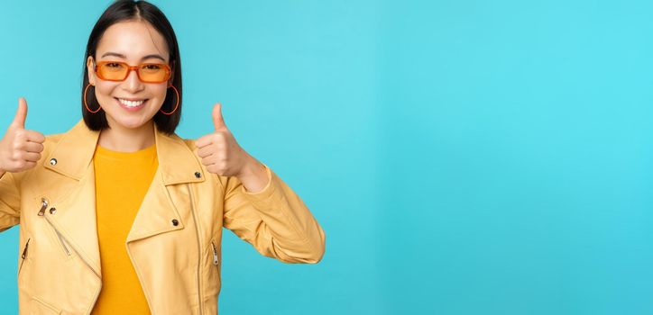 Portrait of modern young asian chinese woman in sunglasses, wears trendy outerwear, showing thumbs up and smiling pleased, recommending smth, blue background.