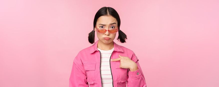 Portrait of asian girl looks confused and points at herself, perplexed face, stares with disbelief at camera, stands over pink background.