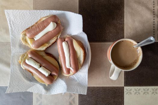 russian hot sandwiches with cheese, sausages and mayonnaise with cup of coffee with milk on table with square pattern, bachelor breakfast concept, high angle view