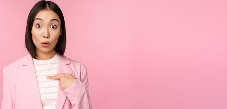 Portrait of asian businesswoman reacts surprised, points at herself with disbelief on face, posing in suit against pink background.