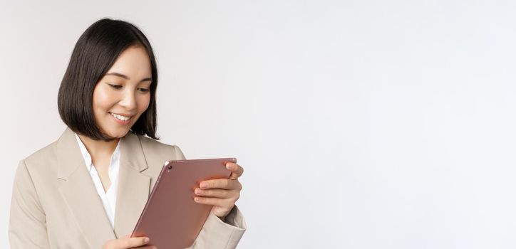 Image of asian businesswoman using digital tablet, looking at gadget and smiling, working, standing against white background.