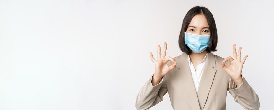Coronavirus and workplace concept. Image of asian saleswoman, company worker in medical mask showing okay sign, smiling pleased, white background.