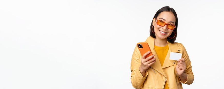 Online shopping and delivery concept. Happy korean girl in stylish clothes, holding credit card and smartphone, laughing and smiling, standing over white background.