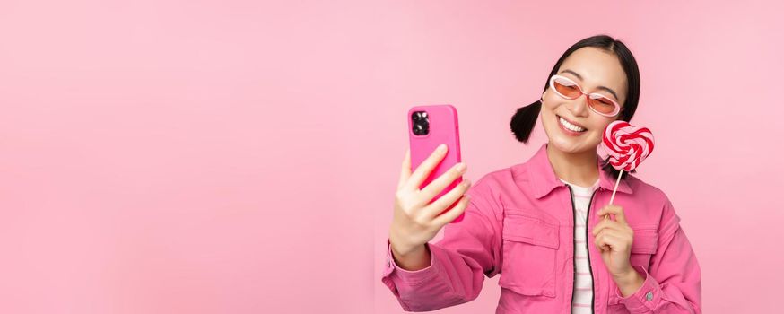 Portrait of stylish, happy asian girl taking selfie with candy, lolipop sweets and smiling, taking photo with mobile app, standing over pink background.