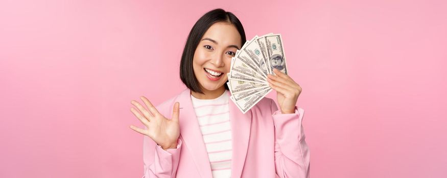 Microcredit, investment and business people concept. Young asian businesswoman, corporate lady showing money, cash dollars, waving hand, pink background.